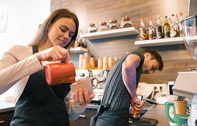 Nuori nainen pit&auml;&auml; kahvikuppia k&auml;dess&auml;&auml;n ja toisella kaataa siihen kahvia. H&auml;nen takanaan nuori mies valmistaa jotakin kahvilan tuotevalikoimaan kuuluvaa tuotetta.